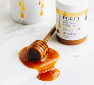 Puriti Premium Pure Raw Manuka Honey UMF 15+ 250g 8.8 oz Jar next to a honey dipper covered in Manuka honey on a white board