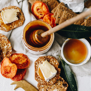 Puriti Premium Raw Manuka Honey UMF 8+ 250g 8.8 oz Jar with a honey dipper in the top of the pot amongst cheese