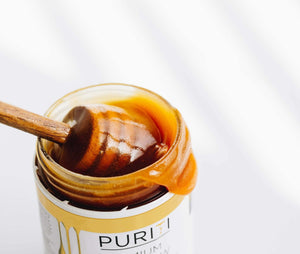 Puriti Premium Manuka Honey Jar with the lid off, honey dipper resting in the top of the jar with Manuka Honey drizzling